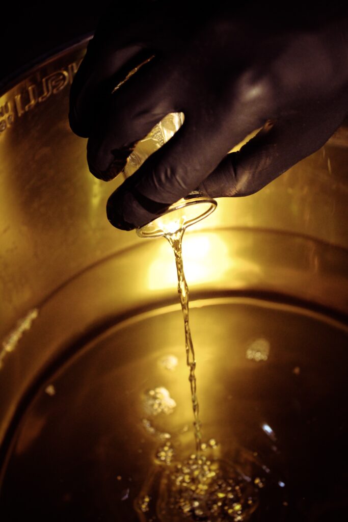 Gloved hand pouring a small scientific glass beaker of liquid into a larger metal vat.